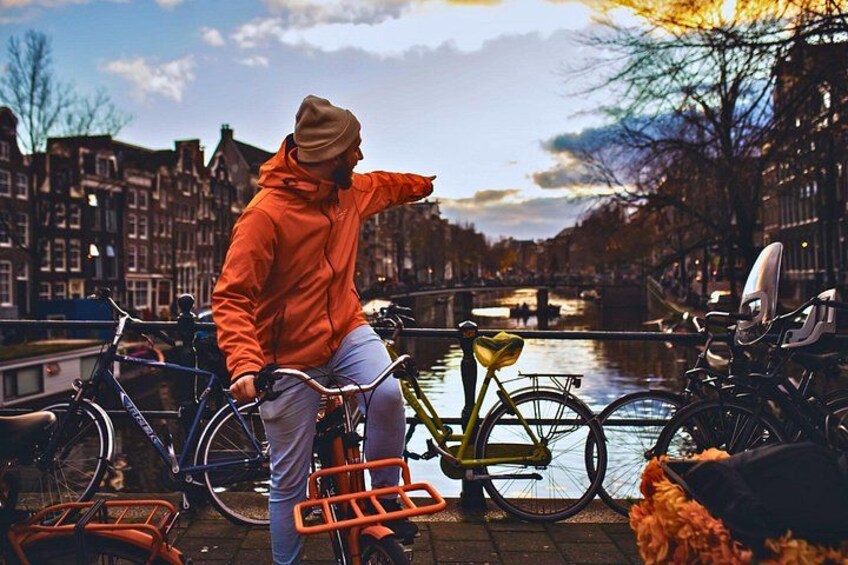 A person on a bicycle with a yellow frame, a red jacket, and a brown beanie, standing by a canal with buildings in the background. 3