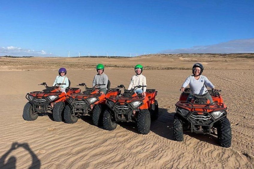 Four people on ATVs in a desert with wind turbines in the background. 1