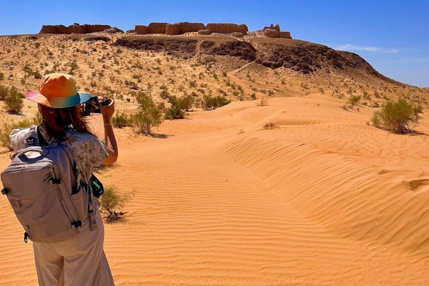 A person taking a photo of a desert landscape with sand dunes and a distant rocky formation. 3
