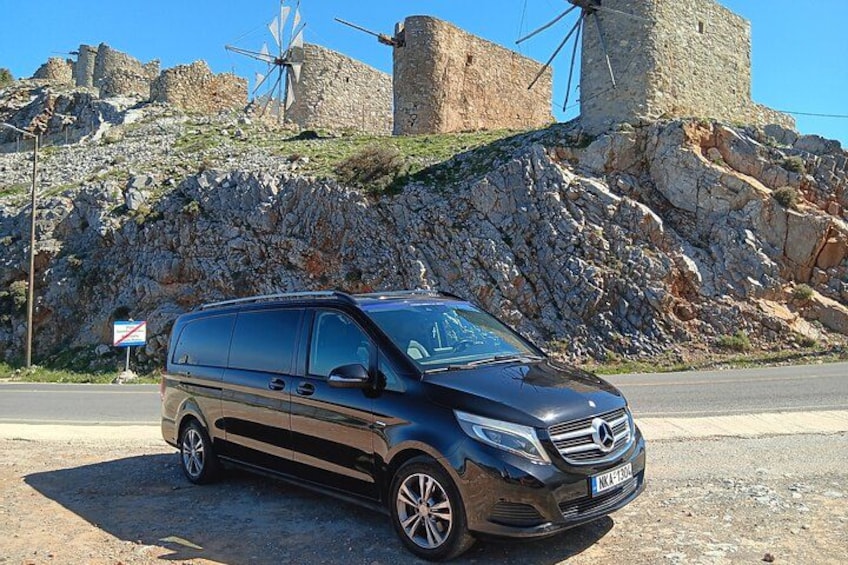 Un fourgon noir de marque Mercedes-Benz était garé sur une route, avec un vieux moulin à vent et des murs de pierre en arrière-plan. 2