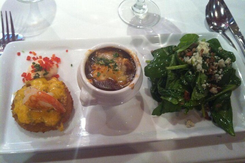 Une assiette rectangulaire blanche sur laquelle sont disposés trois plats : un tartare de crevettes, un bol de soupe et une salade d’épinards. 2