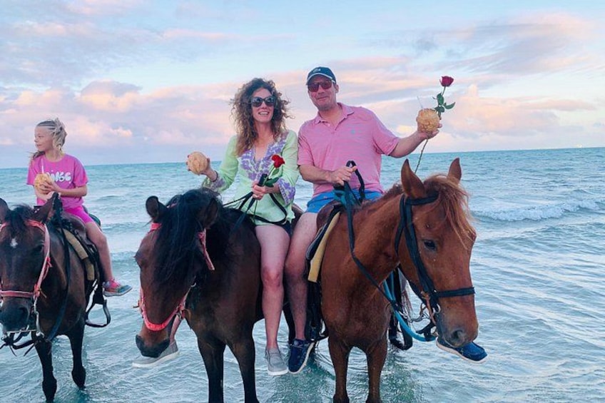 Drie personen rijden op paarden aan het strand, met de oceaan op de achtergrond. 2