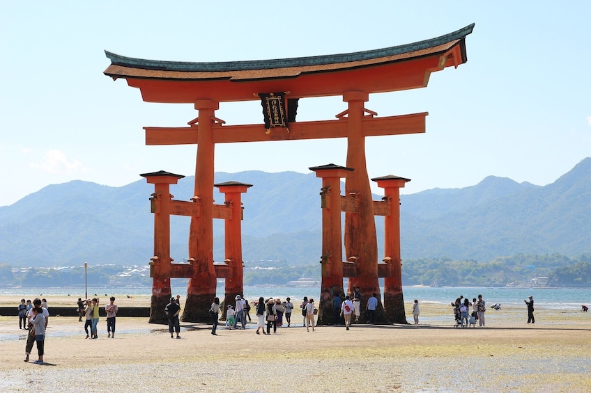 A large red torii gate with a curved roof, surrounded by people and a beach with mountains in the background. 3
