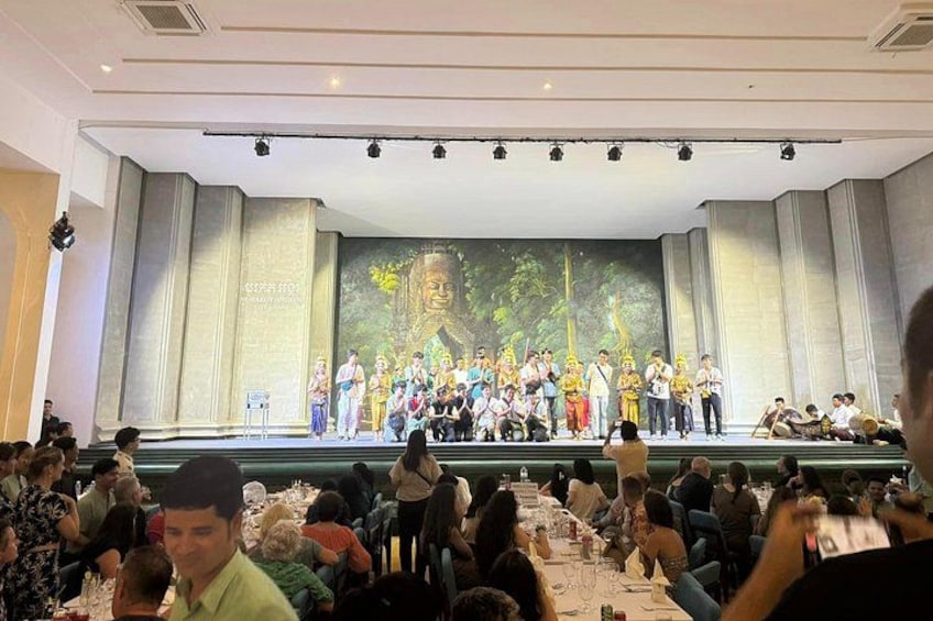 Un escenario con un grupo de personas vestidas con atuendos tradicionales, un público sentado y un telón de fondo con una escena de bosque. 3