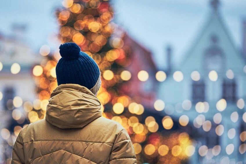 A person from behind wearing a blue beanie and a beige jacket, standing in front of a decorated Christmas tree with lights. 3