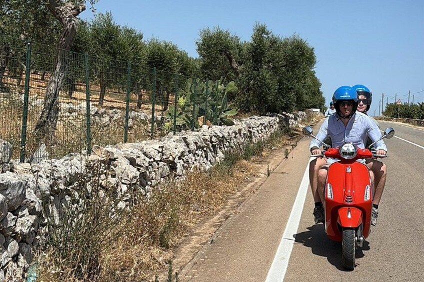 Een rode scooter op een verharde weg, met een stenen muur en begroeiing aan de zijkant. 1