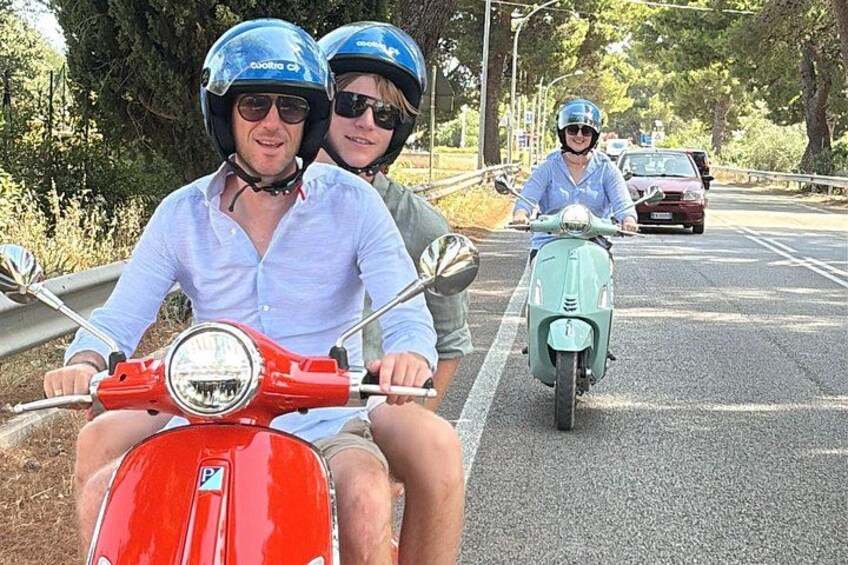 Twee personen op een rode scooter en nog iemand op een groene scooter, op een weg met bomen en een auto op de achtergrond. 4