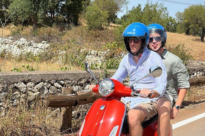 Twee personen op een rode scooter, een stenen muur en een landelijk landschap met bomen. 3