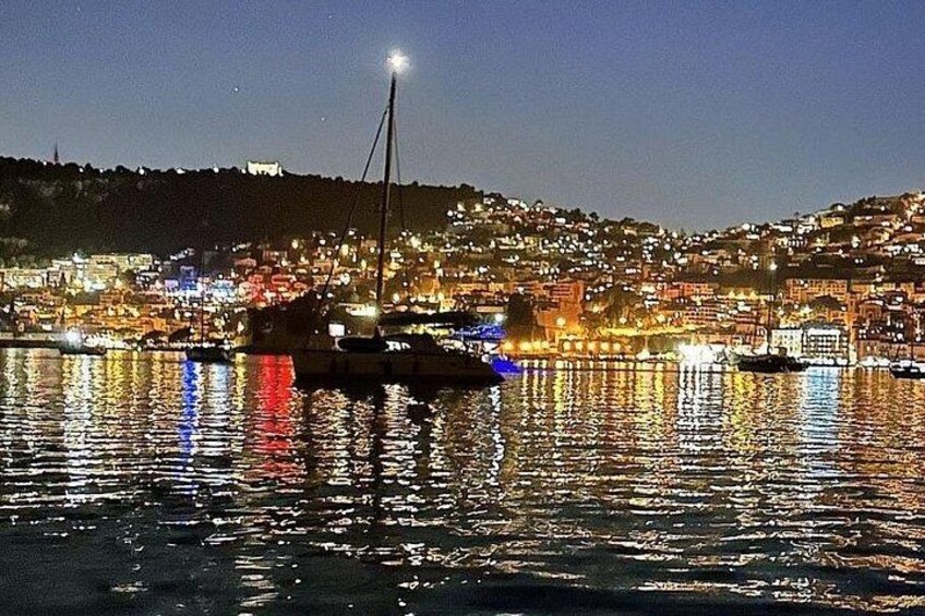 Un port illuminé la nuit, avec des bateaux, une ville perchée sur une colline et un phare imposant. 5