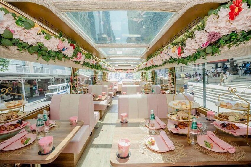 En restaurangvagn på ett tåg med rosa och vit inredning, blomsterarrangemang och bord som är dukade med mat och dryck. 3