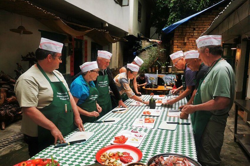 A group of people wearing aprons and chef hats, preparing food at an outdoor table with a checkered tablecloth. 1