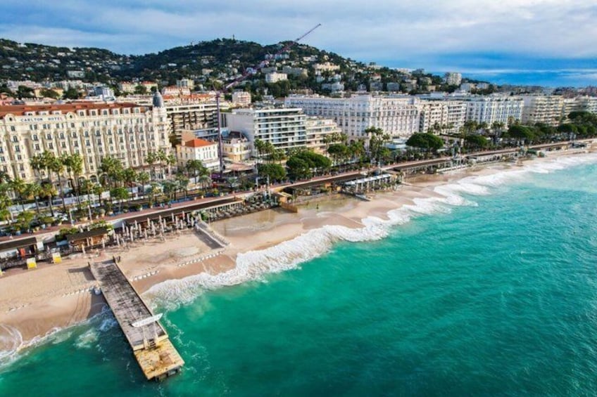 A coastal cityscape with a pier extending into turquoise waters, beachfront buildings, and a hilly backdrop. 1