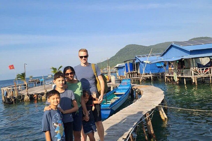 Una familia en un muelle de madera con un bote azul, rodeada de casas tradicionales sobre el agua. 2