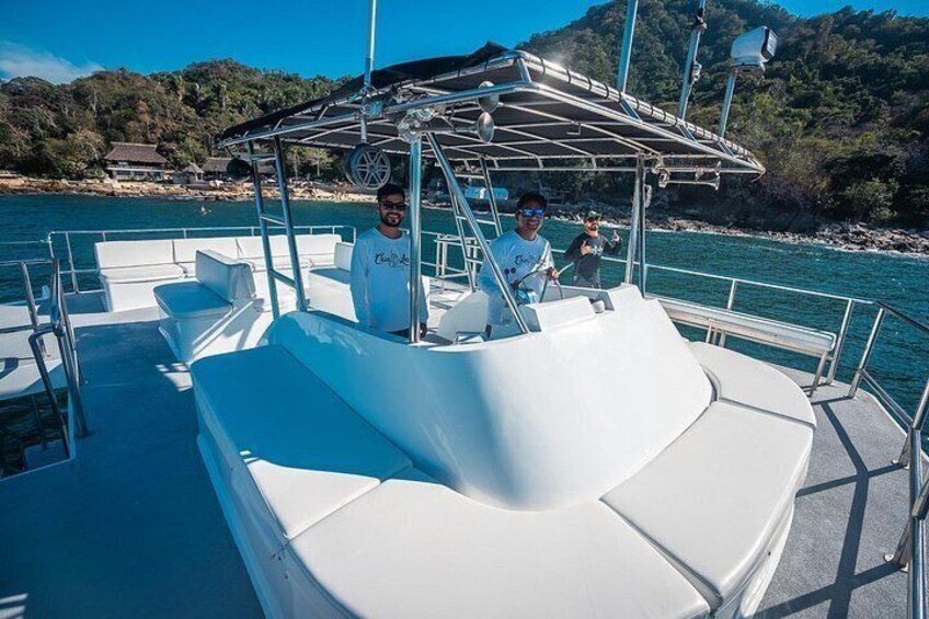 A boat with white seating and a canopy, docked near a coastline with trees and buildings in the background. 5