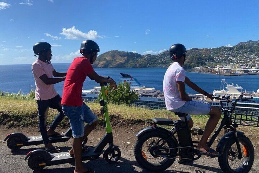 Three people riding electric scooters along a coastal road with a view of the ocean and hills in the background. 2