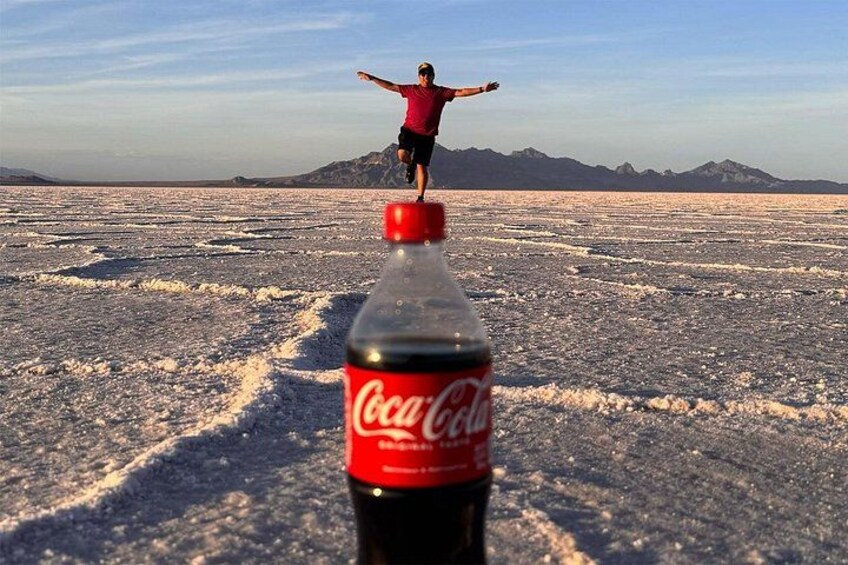 Una persona equilibrándose sobre una botella de Coca-Cola en un desierto de sal, con montañas al fondo. 4