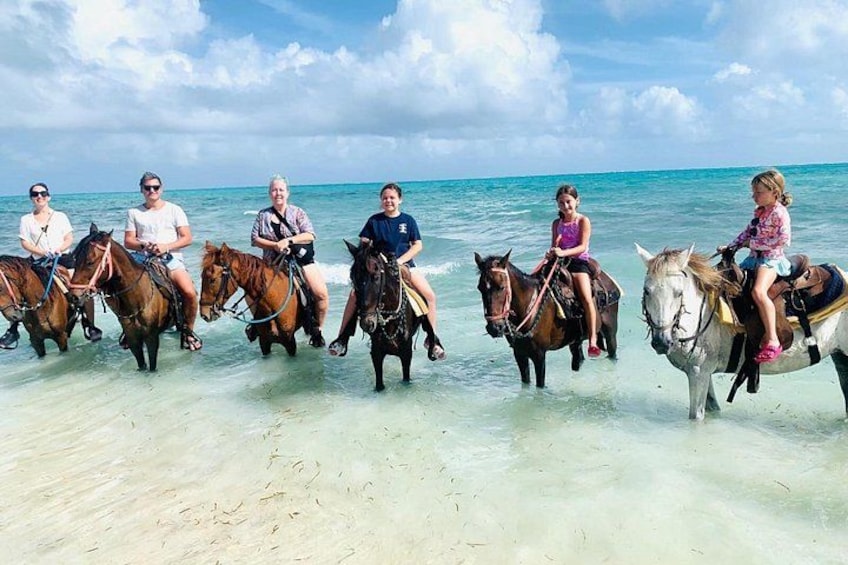A group of people riding horses on a beach with clear water and a cloudy sky. 2