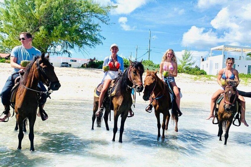 People riding horses on a beach with clear water and a white building in the background. 3