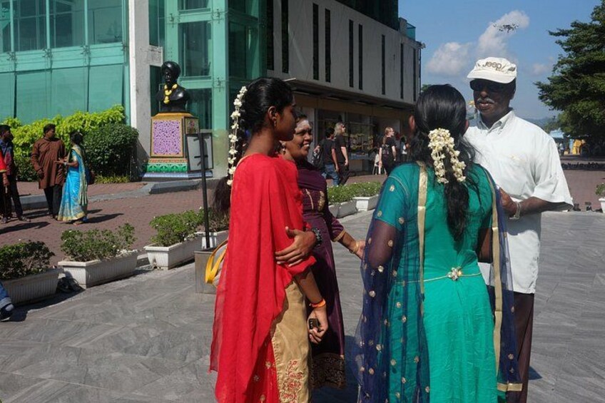 Eine Gruppe von Menschen in traditioneller Kleidung steht auf einem öffentlichen Platz mit einer Statue und modernen Gebäuden im Hintergrund. 2