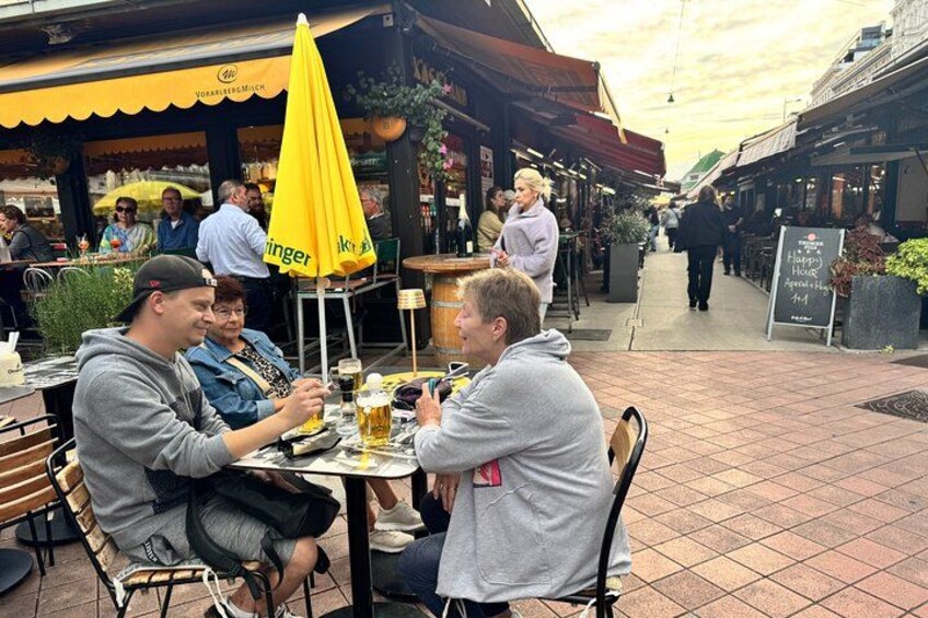 Des personnes sont assises à une table à l’extérieur, devant un marché, et sirotent des boissons à l’ombre d’un parasol jaune. 3