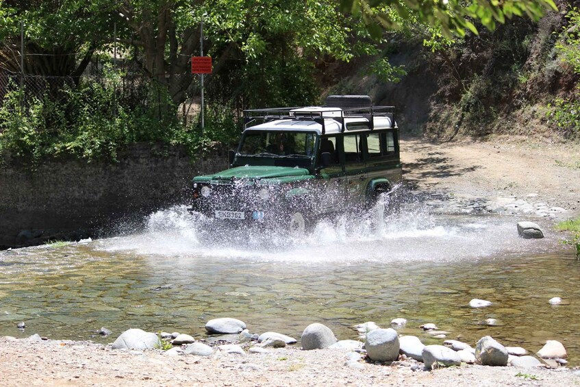 A green off-road vehicle driving through a shallow stream surrounded by rocks and vegetation. 1