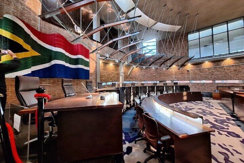 Un salón de conferencias con una gran bandera de Sudáfrica, una mesa larga y varias sillas. 1