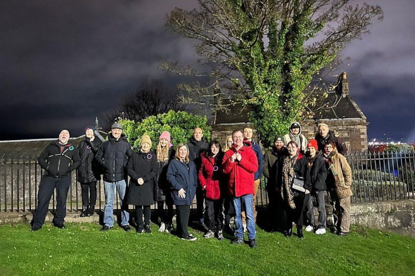 A group of people standing on a grassy area with a stone building and a tree in the background. 1