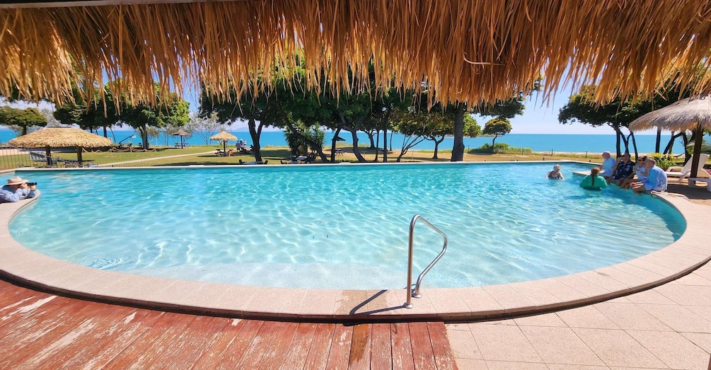 A clear swimming pool with a wooden deck, surrounded by trees and a view of the sea. 2
