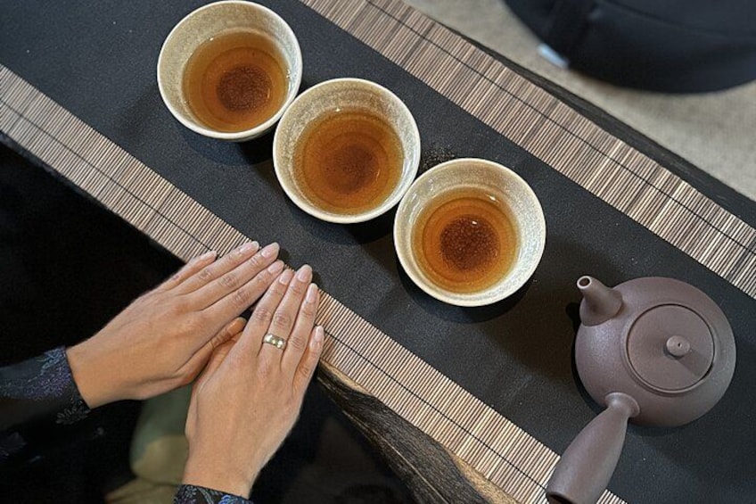 Une personne applaudit en frappant dans ses mains au-dessus de trois bols de thé placés sur une table, à côté d’une théière. 2