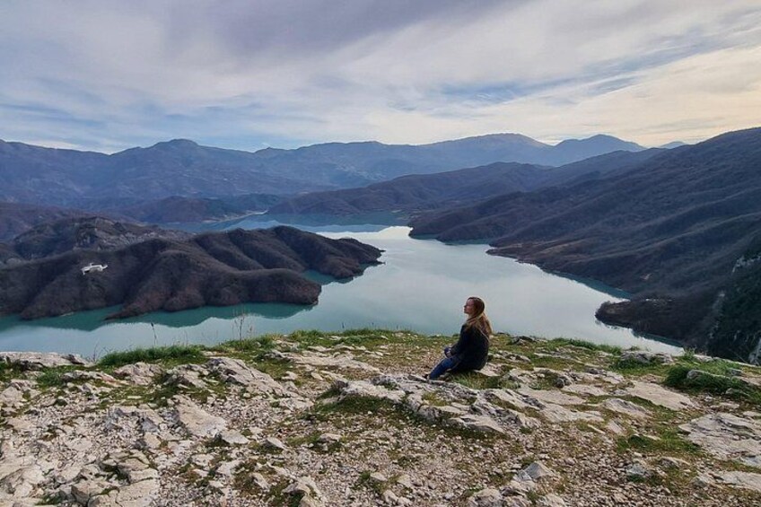 A person sitting on a rocky outcrop overlooking a valley with a lake and mountains. 4