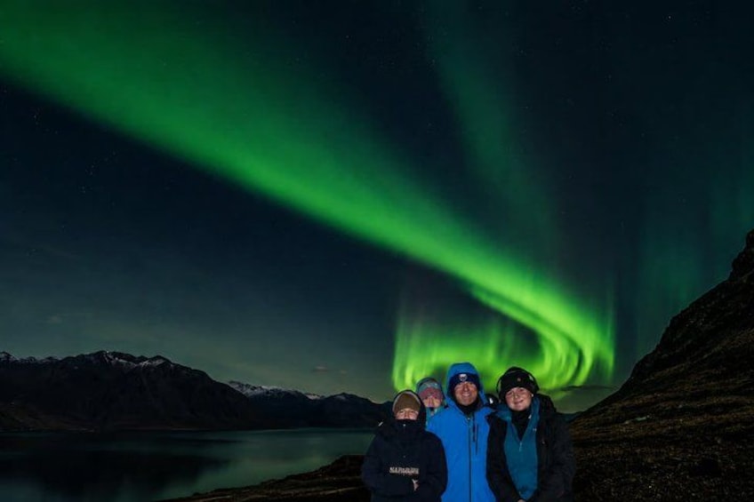 Un grupo de personas de pie cerca de un lago, con una cordillera al fondo y las auroras boreales en el cielo. 3