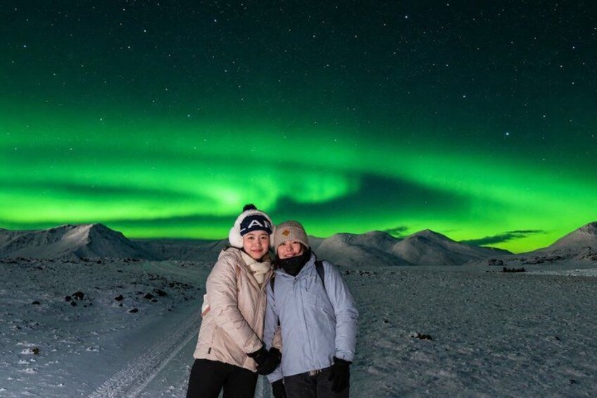 Two people standing on a snow-covered path with a green aurora in the background. 1
