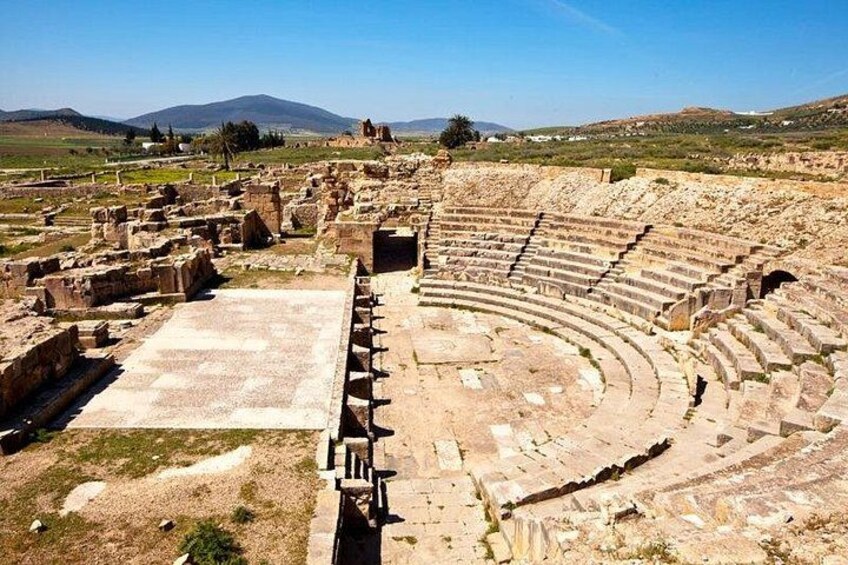 Un ancien amphithéâtre doté de gradins en pierre et d’une scène centrale, entouré de ruines et de vastes champs ouverts. 4
