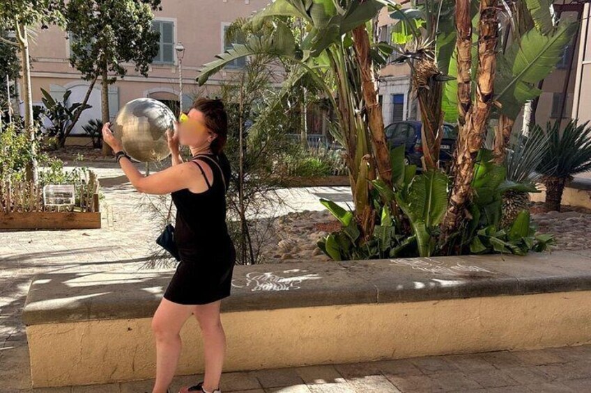 Una persona sostiene una esfera metálica grande, de pie en un jardín con palmeras bananeras y una maceta de concreto. 3
