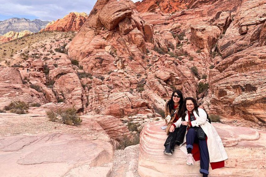 Dos personas sentadas sobre una formación rocosa en un paisaje desértico, con formaciones rojas al fondo. 3