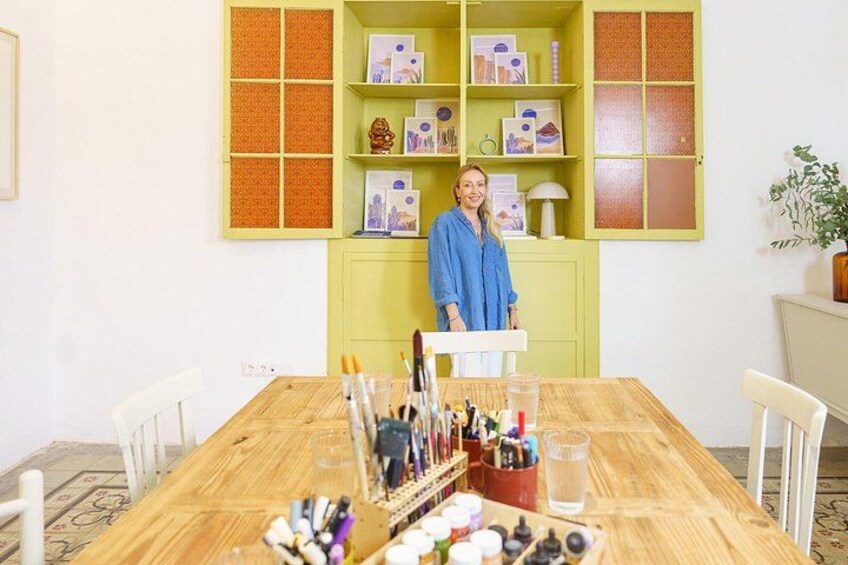 Iemand staat bij een houten tafel met tekensupplies, een groene boekenkast met ingelijste foto's en een potplant. 3