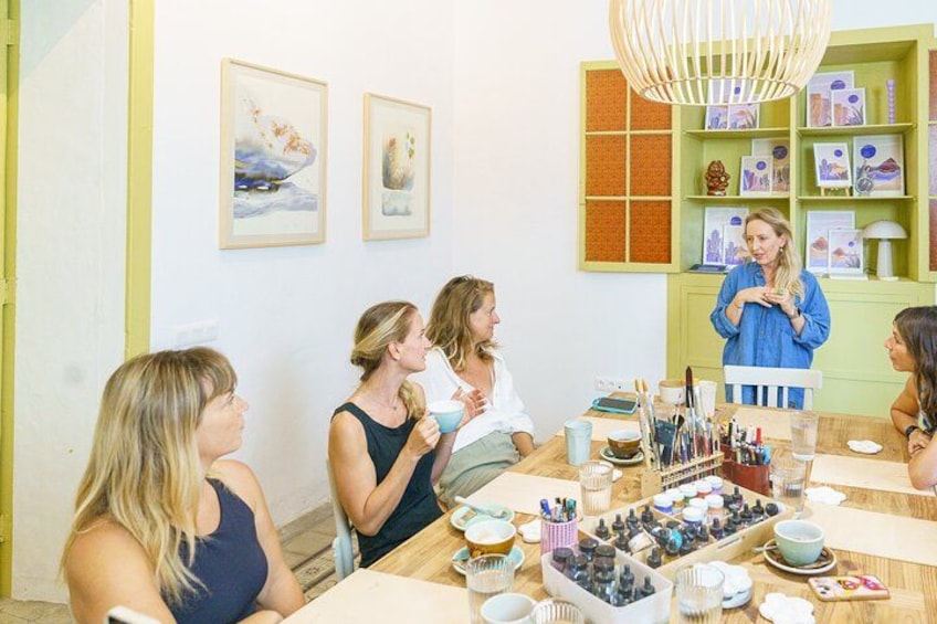 A group of people sitting around a table with various items like cups, bowls, and containers. There are framed pictures on the wall and a green shelf with books and decorative items. 1