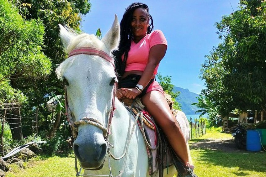 Una persona montando un caballo blanco en un entorno verde y exuberante, con árboles y una montaña al fondo. 3