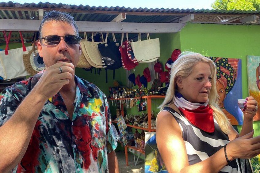A person in a colorful shirt and another in a striped top with a red scarf, standing in front of a market stall with various items and a colorful painting in the background. 3
