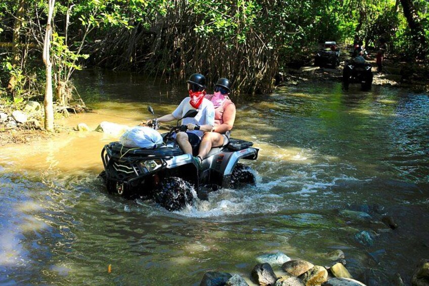 A person riding an ATV through a shallow river surrounded by trees. 1