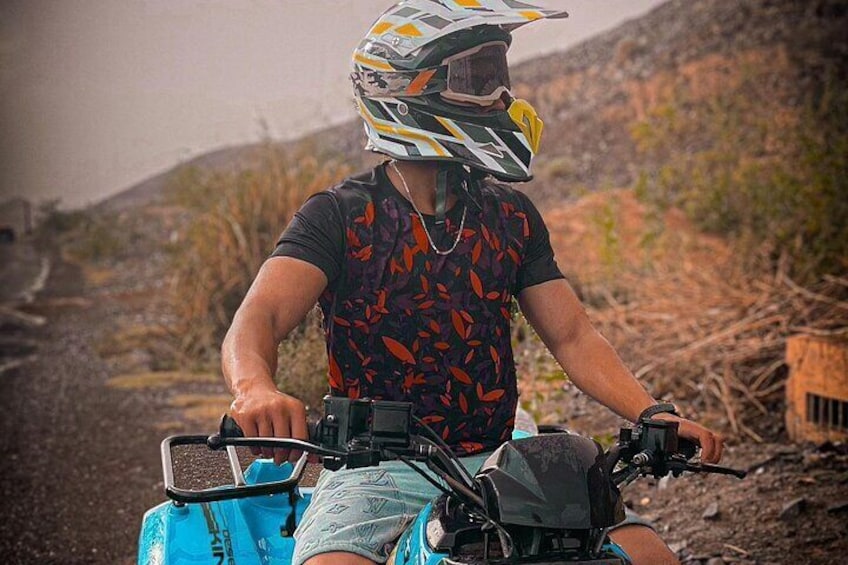 A person riding a blue ATV on a dirt path with sparse vegetation and hills in the background. 3