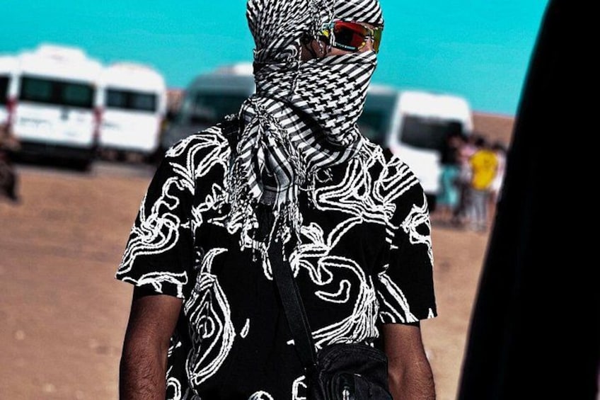 A person wearing a patterned shirt and a headscarf, standing outdoors with vehicles in the background. 5