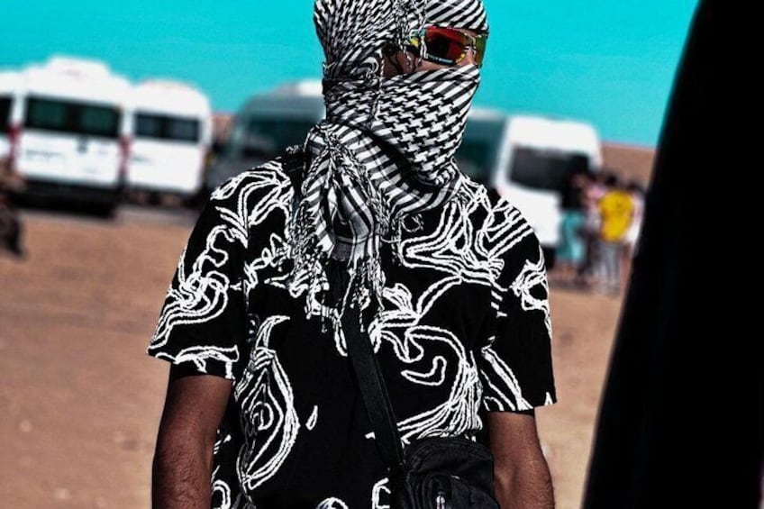 A person wearing a patterned black and white shirt and a headscarf, standing outdoors with vehicles in the background. 2