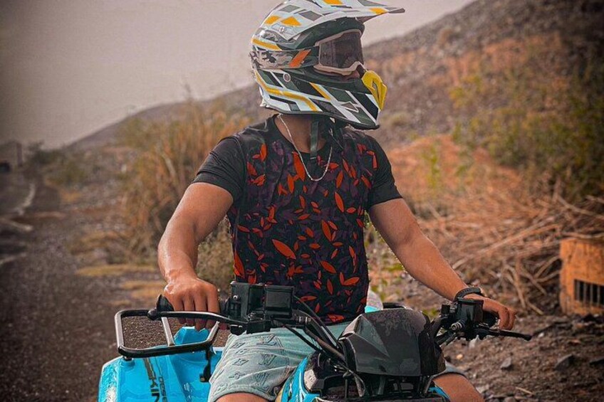 A person riding a blue ATV on a dirt path with sparse vegetation and hills in the background. 2