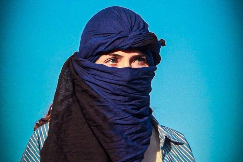 A person wearing a blue headscarf and a striped shirt against a blue background. 5