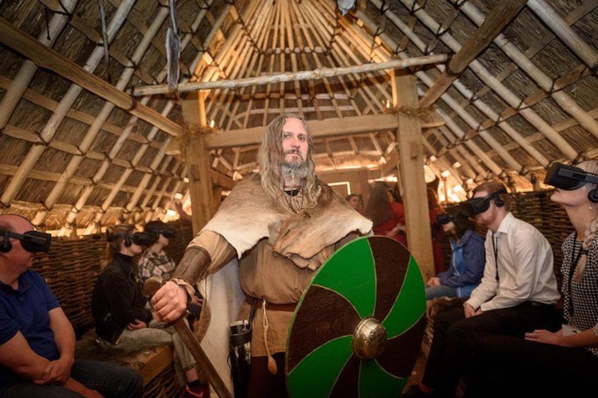 A person in a historical costume holding a shield, surrounded by people in a thatched-roof structure. 1