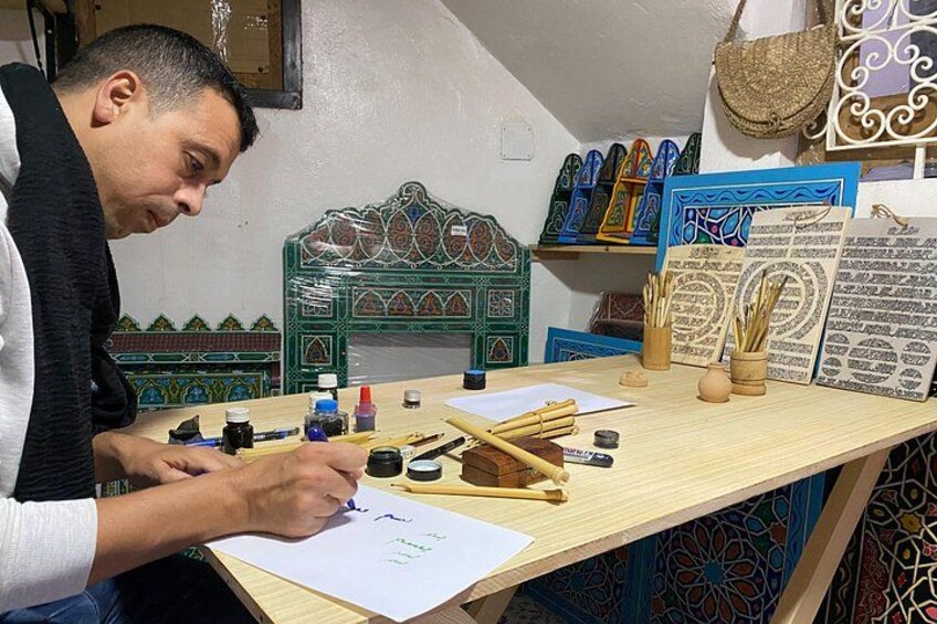 Una persona escribiendo en papel con herramientas de caligrafía, rodeada de baldosas decorativas y una pared con diseño. 3