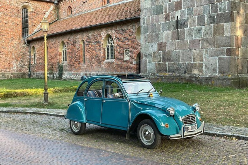 A teal vintage car parked on a cobblestone street in front of a brick building with arched windows. 1