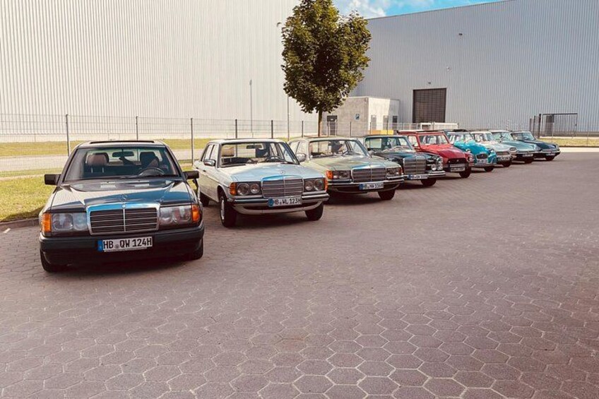 A row of vintage cars parked in a lot with a large industrial building in the background. 2