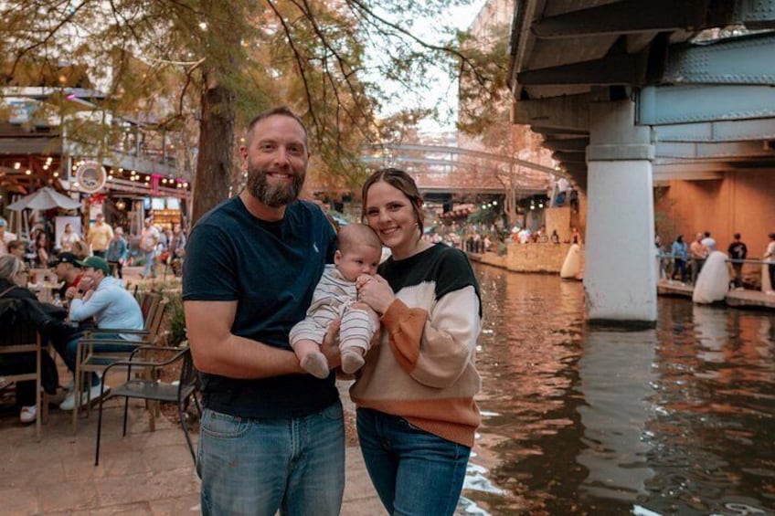 A couple holding a baby by a riverside with people and outdoor seating in the background. 5